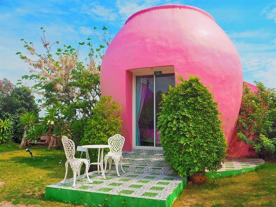 粉色水缸造型小屋，屋前摆着白色桌椅，掩映在花园与晴空之下